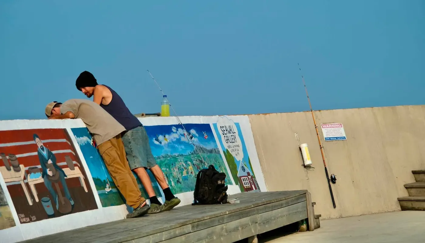 Two people leaning over a painted seawall beside fishing rods in Gimli